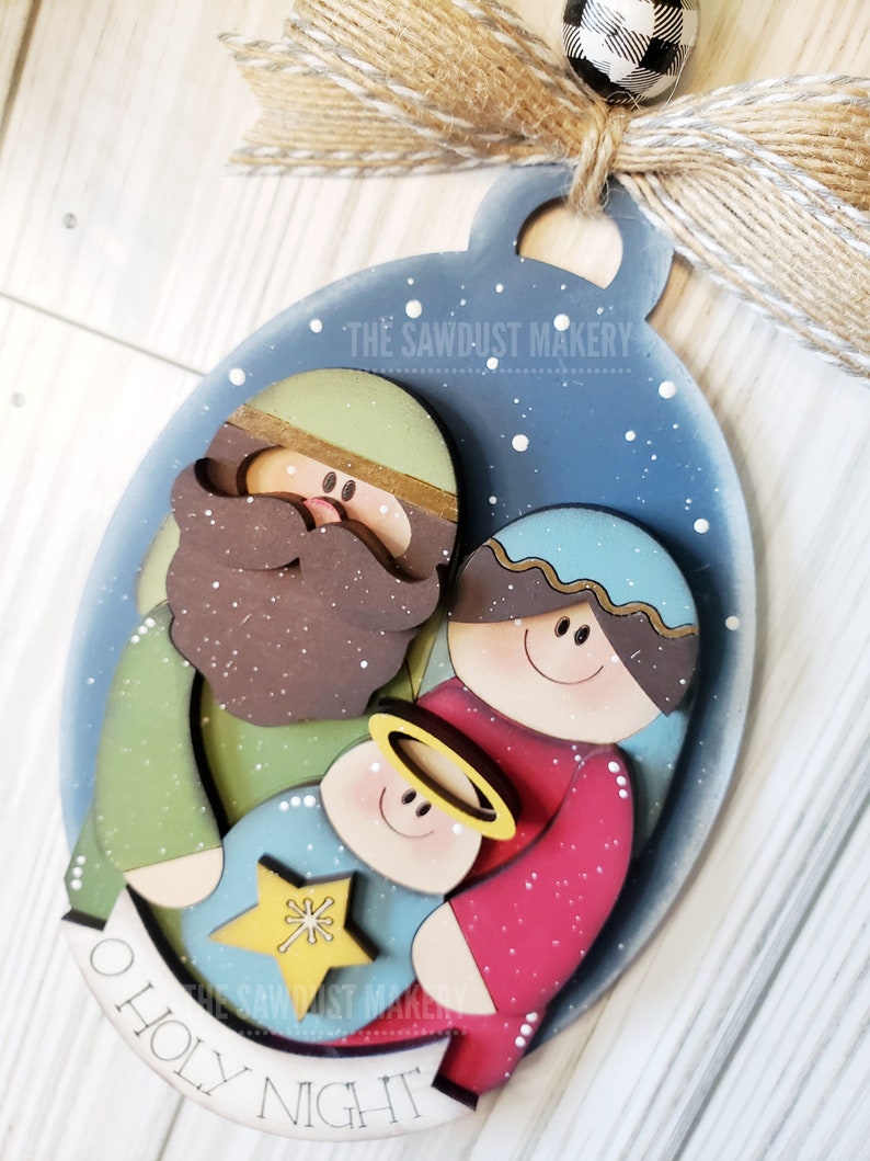 May include: A wooden Christmas ornament depicting the nativity scene with Joseph, Mary, and baby Jesus. The ornament is painted in a blue, green, red, and brown color scheme and features a white star and the text "O Holy Night".