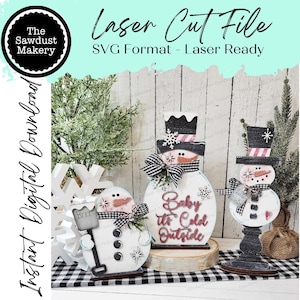 May include: Three wooden snowman figures with black and white paint, one with a red hat and the text "Baby It's Cold Outside", one with a black hat and the text "Brrr", and one with a black hat and a red heart. The snowmen are standing on a black and white checkered surface.