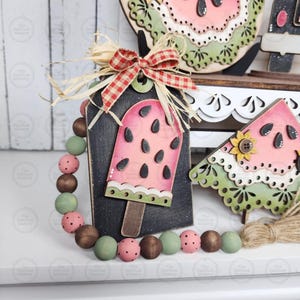 May include: A decorative wooden tag with a watermelon popsicle design, accented with a red and white checkered bow and a beaded garland. The popsicle is pink with black seeds and a wooden stick. The garland features brown, green, and pink beads.