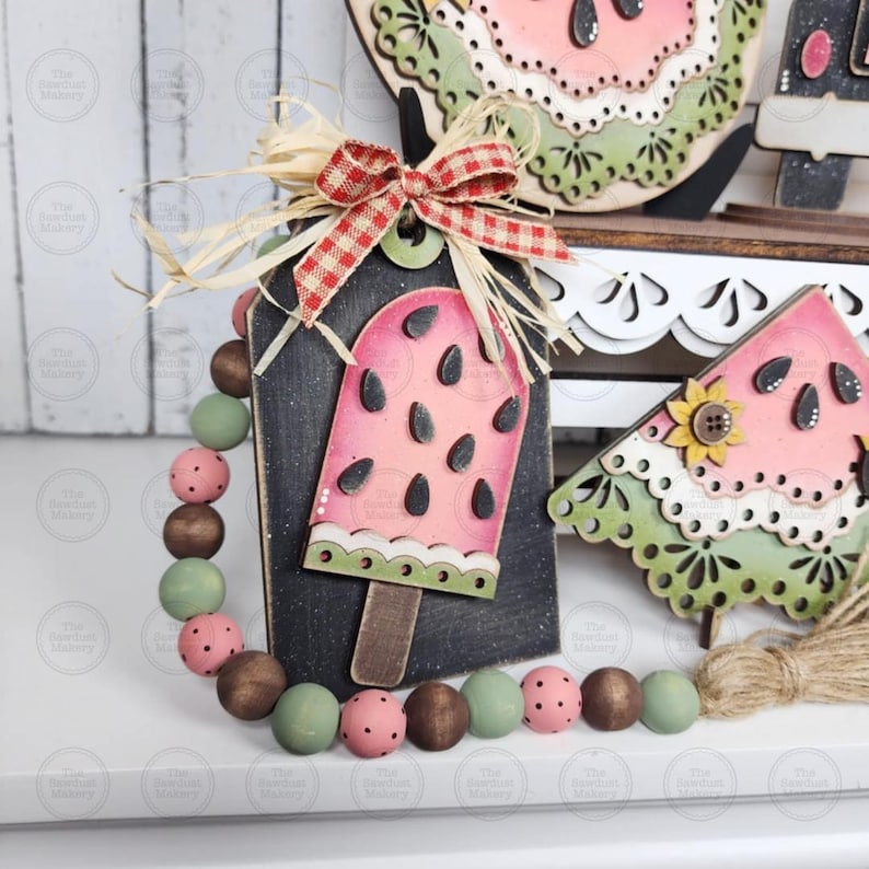 May include: A decorative wooden tag with a watermelon popsicle design, accented with a red and white checkered bow and a beaded garland. The popsicle is pink with black seeds and a wooden stick. The garland features brown, green, and pink beads.