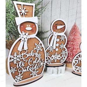 May include: Three wooden gingerbread snowman ornaments with white details. The largest snowman has the text "Baby It's Cold Outside" on its body.