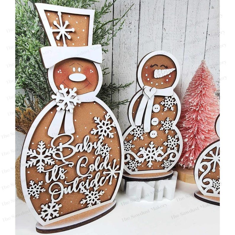 May include: Three wooden gingerbread snowman ornaments with white details. The largest snowman has the text "Baby It's Cold Outside" on its body.