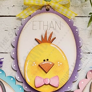 May include: Purple and white scalloped wooden egg with a yellow chick face and a pink bow. The egg has the name "ETHAN" written on it.