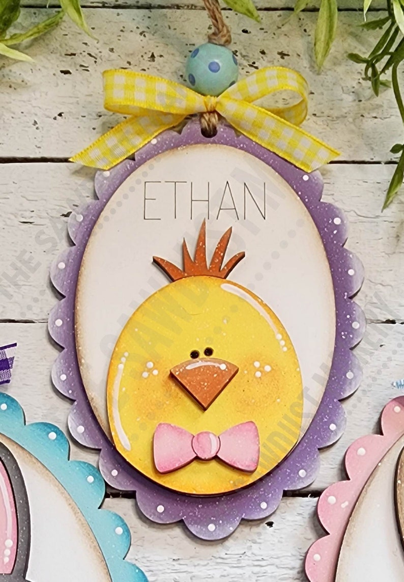 May include: Purple and white scalloped wooden egg with a yellow chick face and a pink bow. The egg has the name "ETHAN" written on it.