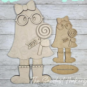 May include: Two unfinished light-colored wood ghost cutouts. Each ghost features a bow, round glasses, a lollipop, and a tag with the word BOO. The larger ghost has legs and boots, while the smaller ghost includes a separate oval wooden base.