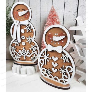 May include: Two wooden gingerbread snowman ornaments with white paint and brown accents. The snowman on the left has a white scarf and snowflakes. The snowman on the right has a white bow tie and hearts.