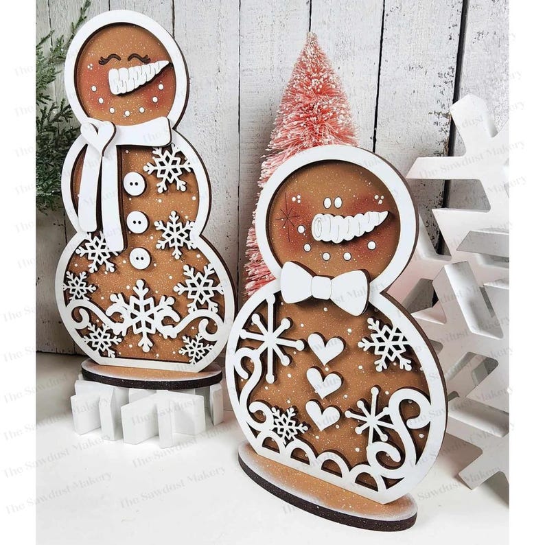 May include: Two wooden gingerbread snowman ornaments with white paint and brown accents. The snowman on the left has a white scarf and snowflakes. The snowman on the right has a white bow tie and hearts.