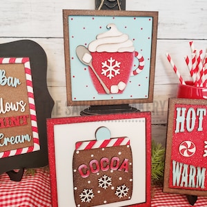 May include: Two wooden signs with a red and white checkered background. The first sign has a red mug with white whipped cream and a snowflake on it. The second sign has a brown cocoa container with white snowflakes on it. Both signs have the words "Hot" and "Warm" on them.