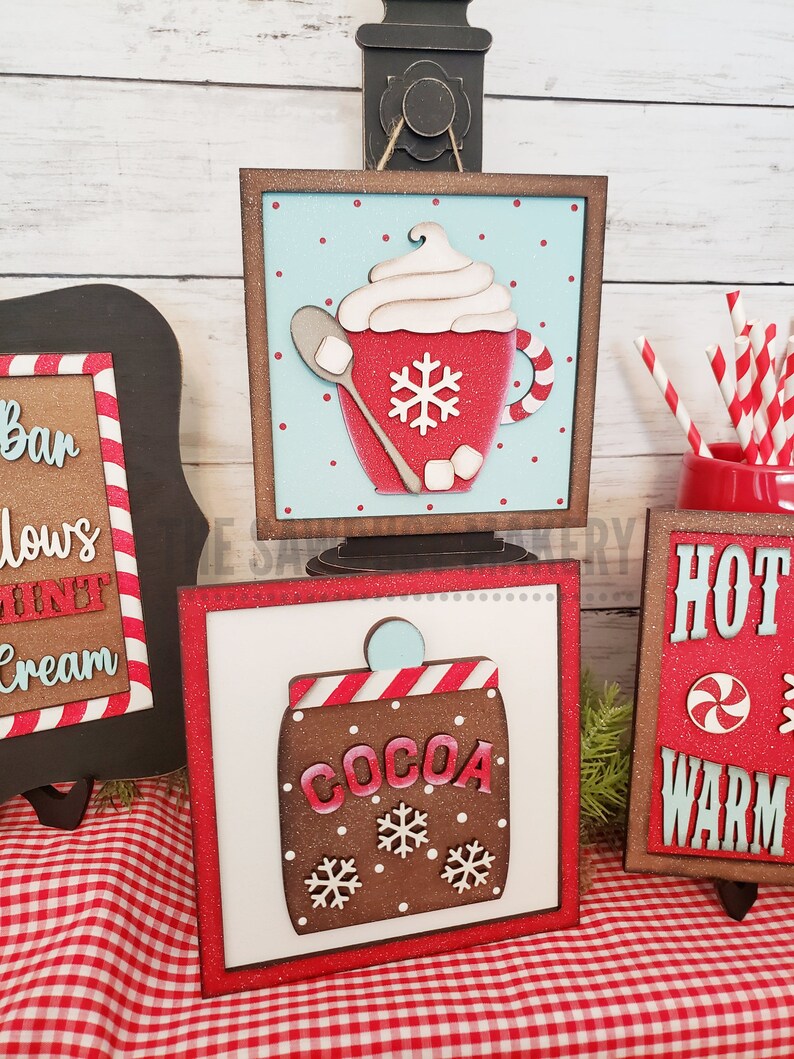 May include: Two wooden signs with a red and white checkered background. The first sign has a red mug with white whipped cream and a snowflake on it. The second sign has a brown cocoa container with white snowflakes on it. Both signs have the words "Hot" and "Warm" on them.