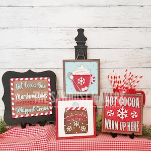 May include: Four wooden signs with a red and white gingham tablecloth in the background. The signs are decorated with a hot cocoa theme and include the text "Hot Cocoa Bar", "Marshmallows", "Peppermint", "Whipped Cream", "Cocoa", "Hot Cocoa", and "Warm Up Here".