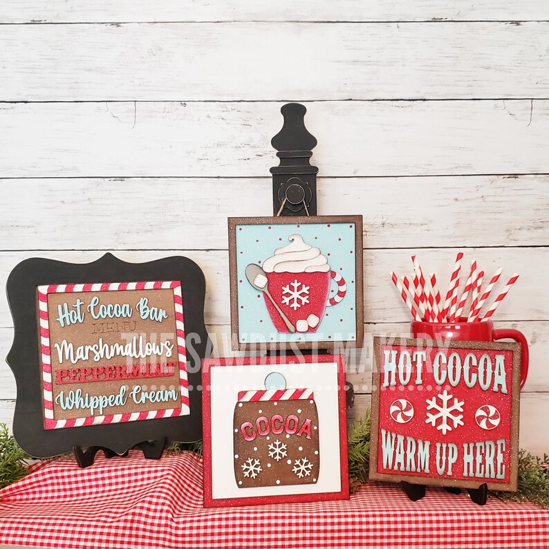 May include: Four wooden signs with a red and white gingham tablecloth in the background. The signs are decorated with a hot cocoa theme and include the text "Hot Cocoa Bar", "Marshmallows", "Peppermint", "Whipped Cream", "Cocoa", "Hot Cocoa", and "Warm Up Here".