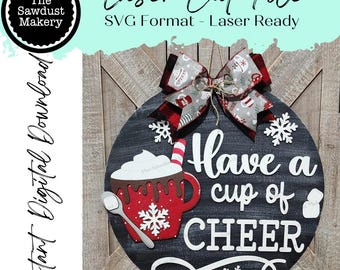 Pode incluir: Um sinal redondo de madeira preto e branco com o texto "Have a cup of CHEER" em branco. O sinal tem uma caneca vermelha e branca com um floco de neve branco e um laço vermelho e branco no topo. O sinal é decorado com flocos de neve brancos.