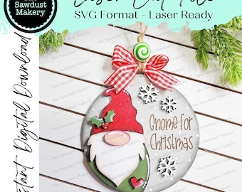 Puede incluir: Un archivo de corte láser para un adorno de gnomo navideño. El gnomo lleva un gorro rojo con un pompón blanco, una barba verde y un corazón rojo en el pecho. El adorno está decorado con copos de nieve y el texto "Gnome for Christmas".