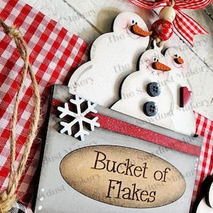 May include: A wooden sign with a red and white checkered background. The sign says "Bucket of Flakes" and has a snowflake and three snowmen on top.