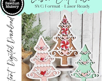 Puede incluir: Tres adornos de árbol de Navidad de madera cortados con láser en rosa, marrón y verde. Cada árbol presenta un borde blanco decorativo, detalles de corazón y una base. La imagen incluye el texto "Laser Cut File" y "Instant Digital Download."