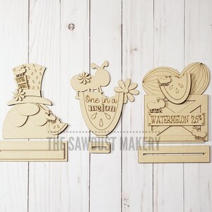May include: Three unfinished wooden cutout signs with watermelon themes. The first sign features a watermelon-hatted character with the text "You're Sweet". The second sign features a watermelon slice with the text "One in a Melon". The third sign features a watermelon slice in a crate with the text "Local Watermelon 25&cent;".