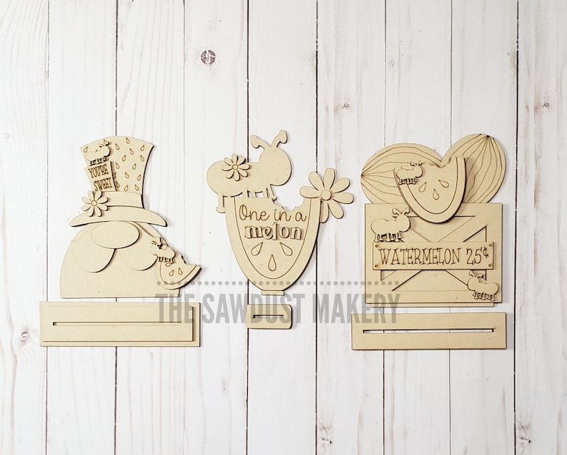 May include: Three unfinished wooden cutout signs with watermelon themes. The first sign features a watermelon-hatted character with the text "You're Sweet". The second sign features a watermelon slice with the text "One in a Melon". The third sign features a watermelon slice in a crate with the text "Local Watermelon 25&cent;".
