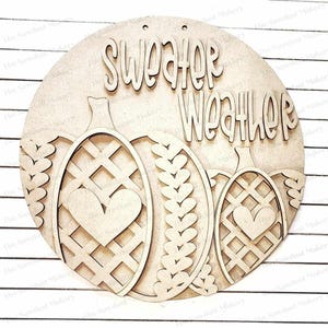 May include: A circular wooden sign with the words "Sweater Weather" in cursive. The sign features three pumpkin shapes with heart and lattice designs. The sign is made of unfinished wood and has two small holes at the top for hanging.