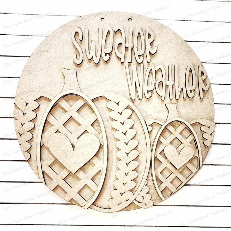 May include: A circular wooden sign with the words "Sweater Weather" in cursive. The sign features three pumpkin shapes with heart and lattice designs. The sign is made of unfinished wood and has two small holes at the top for hanging.