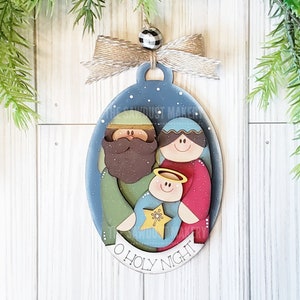 May include: A wooden Christmas ornament featuring a blue and white oval with a depiction of the Holy Family. The ornament has a brown, green, and red color scheme and the text "O Holy Night" at the bottom.