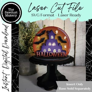 May include: A Halloween-themed laser cut file insert. The design features a purple haunted house with bats and stars against an orange and yellow background. The word "HALLOWEEN" is displayed at the bottom. The insert is on a black stand.