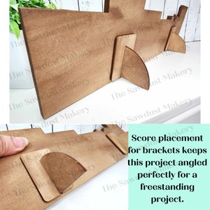 May include: A wooden sign with a brown stain and a score line for folding. The sign is angled to stand upright. The text on the sign reads "Score placement for brackets keeps this project angled perfectly for a freestanding project."
