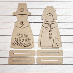 May include: A wooden craft kit featuring a pilgrim couple with the words "Give Thanks" on the man's outfit. The kit includes four separate pieces for the couple's bodies and a base.