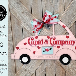 以下が含まれることがあります： ピンクの車の形をした木製の切り抜きで、「Cupid & Company」という文字と、赤と白のチェック柄のリボンが上に乗っています。車は白い水玉模様と赤いハートが付いています。
