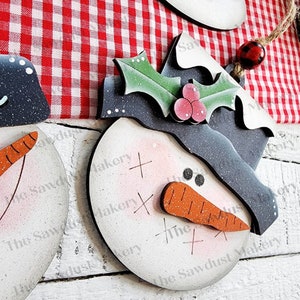 May include: A wooden snowman ornament with a black hat, holly leaves, and red berries. The snowman has a carrot nose, two black eyes, and a white face with a pink blush. The ornament is painted with a distressed white finish.