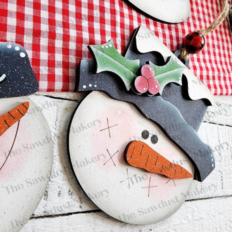 May include: A wooden snowman ornament with a black hat, holly leaves, and red berries. The snowman has a carrot nose, two black eyes, and a white face with a pink blush. The ornament is painted with a distressed white finish.