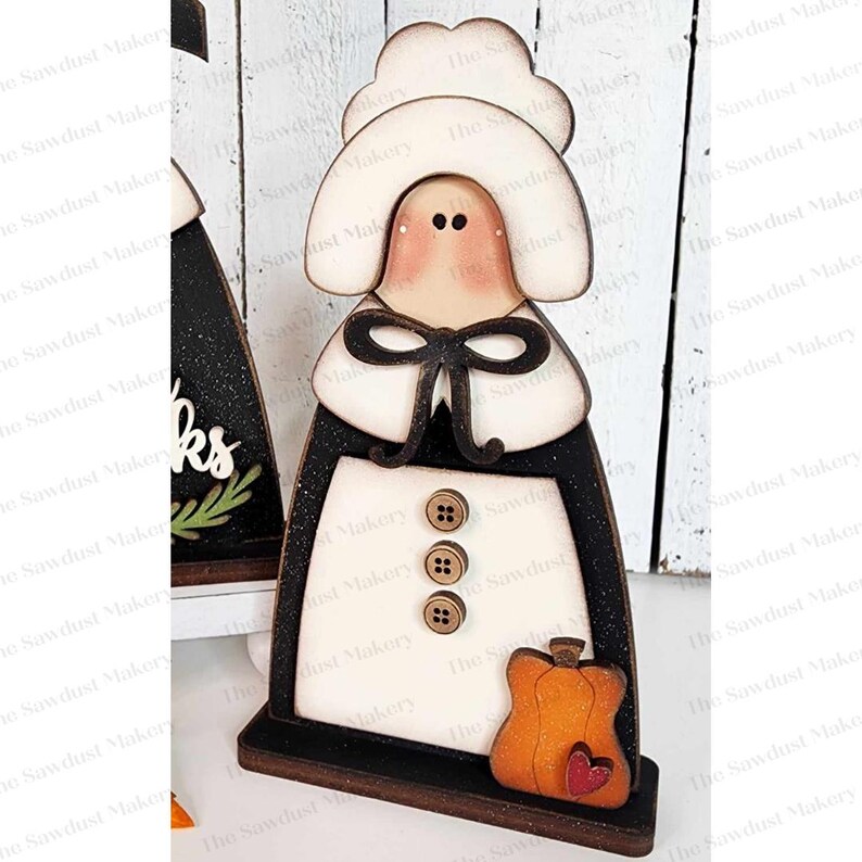 May include: Wooden figurine of a pilgrim woman with a white bonnet, brown dress, and a pumpkin in her hand. The figurine is painted in white, brown, and orange colors. The background is a wooden surface.