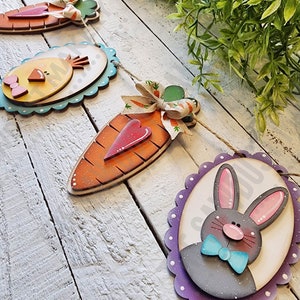 May include: A wooden Easter garland with a chick, carrots, and a bunny. The garland is painted in bright colors and has a scalloped edge.