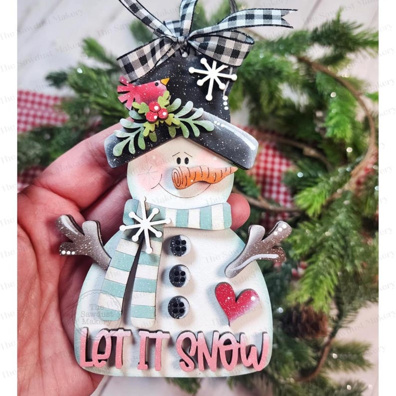 May include: A snowman ornament with a black top hat, a blue and white striped scarf, and a carrot nose. The ornament has a red heart and the words "LET IT SNOW" in pink. A black and white checkered ribbon is at the top.