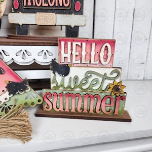 May include: A wooden summer-themed decoration with the words "Hello Sweet Summer" in layered, colorful lettering. The design includes a small bird and a sunflower accent, with a watermelon slice nearby. The base is brown.