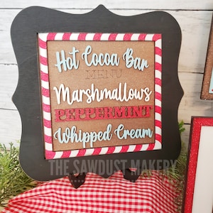 May include: A black framed sign with a red and white candy cane striped border. The sign says "Hot Cocoa Bar Menu" with the words "Marshmallows", "Peppermint", and "Whipped Cream" written in a decorative font. The sign is on a red and white checkered tablecloth.
