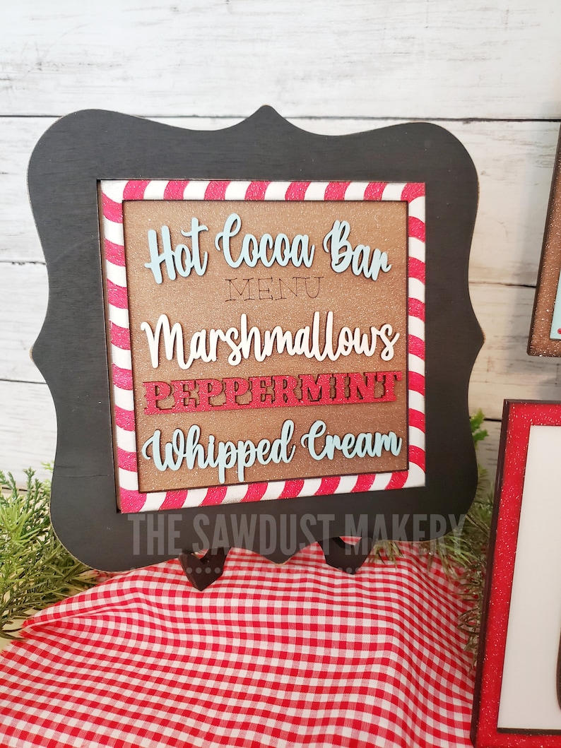 May include: A black framed sign with a red and white candy cane striped border. The sign says "Hot Cocoa Bar Menu" with the words "Marshmallows", "Peppermint", and "Whipped Cream" written in a decorative font. The sign is on a red and white checkered tablecloth.