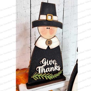 May include: Wooden figurine of a pilgrim with a black hat and white collar. The figurine is holding a sign that says "Give Thanks" with a green leaf garland.