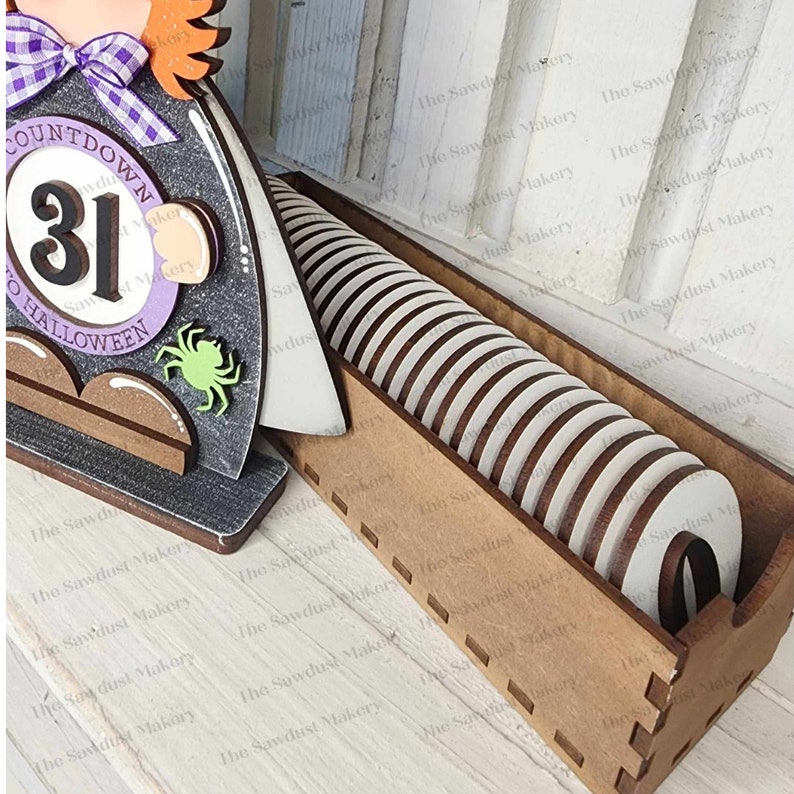 May include: A wooden Halloween countdown calendar with a witch figure and a number 31 on a purple circle. The calendar has a slot for 31 wooden circles to be placed in, with 29 circles already in place.