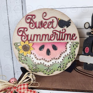 May include: Wooden round sign with the words "Sweet Summertime" in red script. The sign features a watermelon slice, a sunflower, and a small black bird. The sign is a summer-themed decoration.
