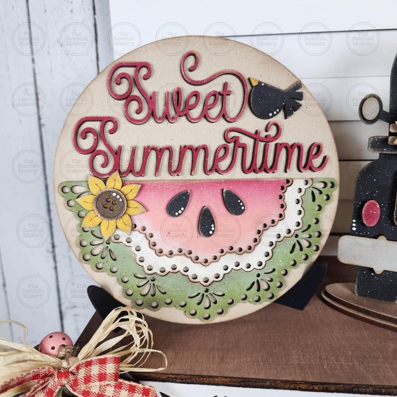 May include: Wooden round sign with the words "Sweet Summertime" in red script. The sign features a watermelon slice, a sunflower, and a small black bird. The sign is a summer-themed decoration.