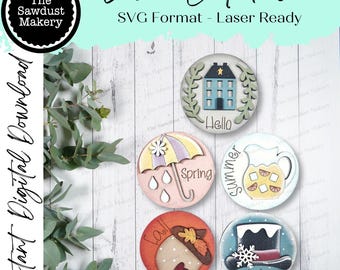 May include: Five round wooden signs with different seasonal themes. The signs are painted in various colours and feature illustrations of a house, an umbrella, a pitcher, an acorn, and a hat. The signs also include the words "Hello", "Spring", "Summer", "Fall", and "Winter".