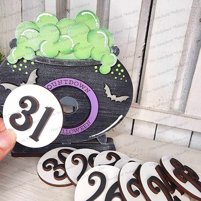 Puede incluir: Un caldero negro con l&iacute;quido verde burbujeante y un anillo morado alrededor de la palabra "COUNTDOWN" y la palabra "HALLOWEEN" en la parte delantera. Hay 8 c&iacute;rculos blancos con n&uacute;meros negros en ellos, incluyendo un c&iacute;rculo con el n&uacute;mero "31" en &eacute;l.