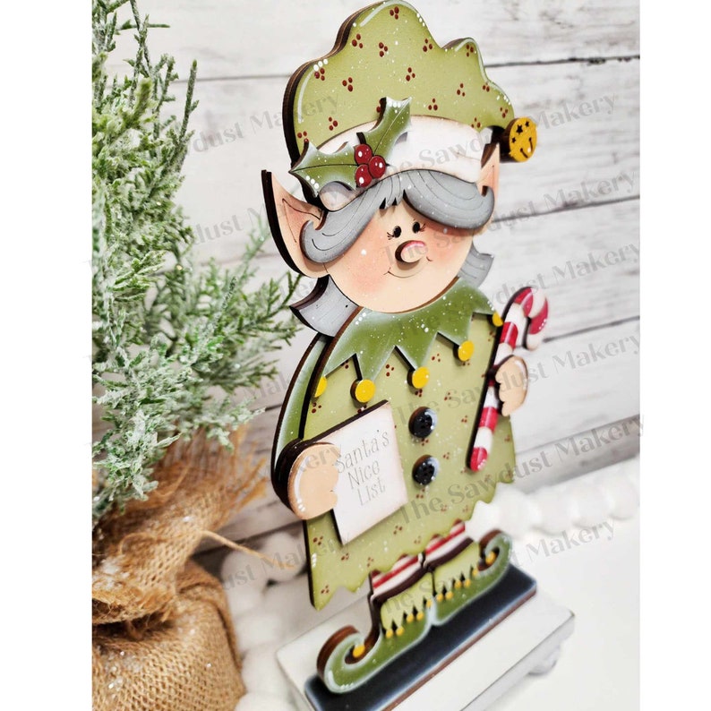 May include: A wooden Christmas elf figurine with a green hat, green and yellow outfit, and a candy cane in its hand. The elf is holding a white paper with the text "Santa's Nice List" written on it.