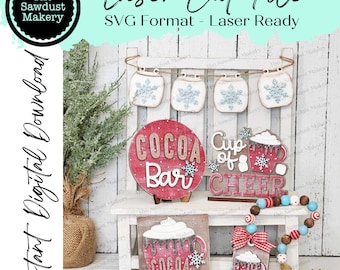 May include: A set of laser cut SVG files for creating a winter-themed cocoa bar sign. The set includes a round sign that says "Cocoa Bar", a square sign that says "Cup of Cheer", a smaller square sign with a mug of cocoa, and a tag with a mug of cocoa. The signs are decorated with snowflakes and red and white accents.
