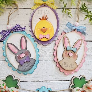 May include: Three wooden Easter egg ornaments with painted animal faces and names. The ornaments are decorated with pastel colors and scalloped edges. The names on the ornaments are Ethan, Chris, and Olivia. A banner with carrots and green leaves hangs below the ornaments.
