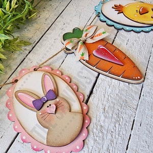 May include: A wooden Easter banner with three hanging decorations. The decorations are a brown bunny with pink ears and a purple bow, an orange carrot with a red heart, and a yellow chick with a pink bow. The banner is strung with twine.