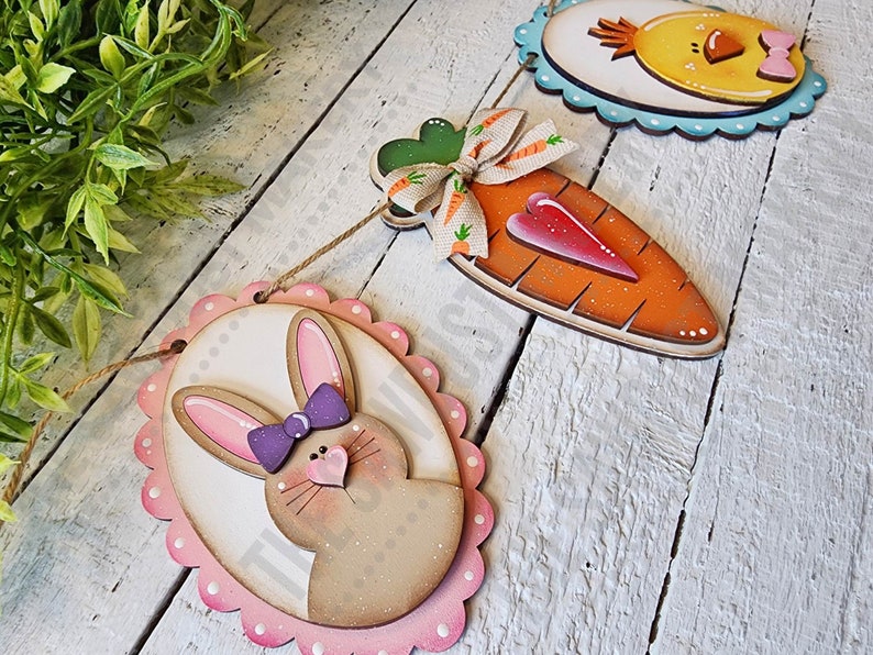 May include: A wooden Easter banner with three hanging decorations. The decorations are a brown bunny with pink ears and a purple bow, an orange carrot with a red heart, and a yellow chick with a pink bow. The banner is strung with twine.