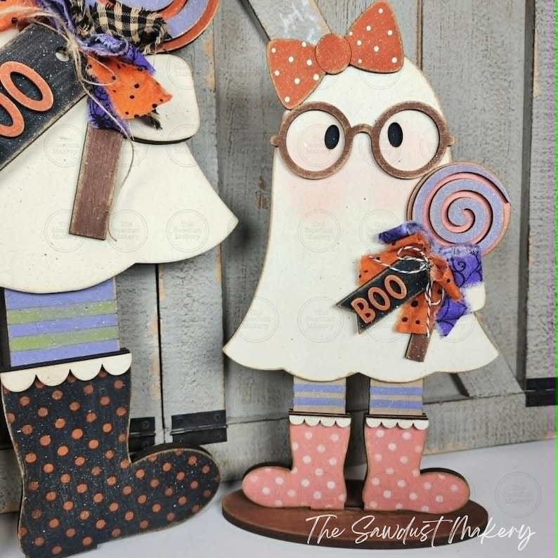 May include: A wooden ghost cutout decoration featuring a white body, round brown glasses, and an orange polka dot bow. The ghost wears pink polka dot boots and holds a purple and orange swirl lollipop with a tag that reads BOO.