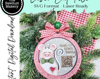 Puede incluir: Adorno navideño con diseño de ratón, corazón rojo y pajarita verde. El adorno incluye el texto "Scan to track Santa" y "'Twas the night before Christmas". Colgando de un árbol de Navidad.
