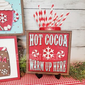 May include: Red wooden sign with white glitter and the text "HOT COCOA WARM UP HERE" in blue lettering. The sign has a white snowflake and two red and white peppermint candies on it. The sign is on a small black easel.